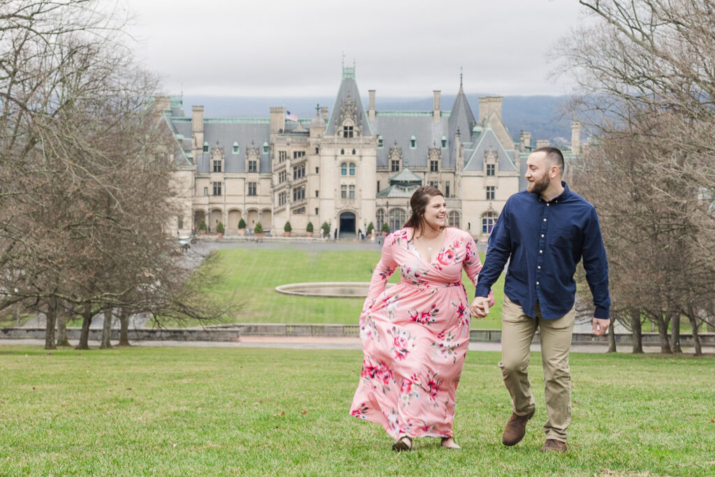 couple holding hands and smiling at each other as they walk to the statue of Diana with the Biltmore Estate in the background