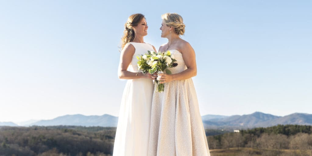 wide image of Brides holding bouquets standing on the south terrace smiling at each other with the blue ridge mountains in the background