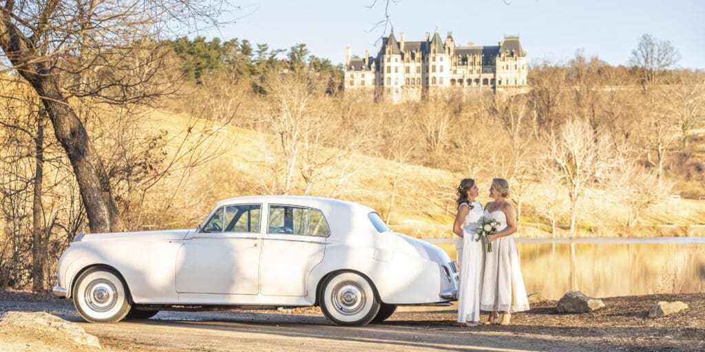 Brides standing by the lagoon with their vintage rolls-royce and Biltmore Estate in the background
