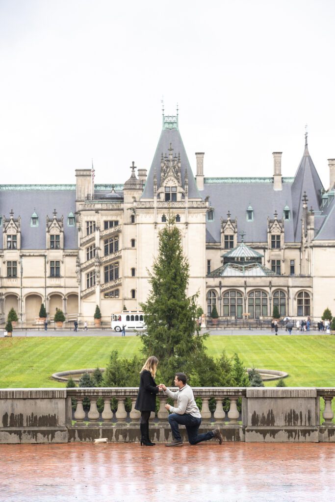 Biltmore estate proposal, man on one knee proposing to his girlfriend on the esplanade with the estate and christmas tree in the background on a rainy day