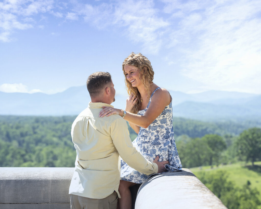 newly engaged couple looking at each other and smiling while she sits on the ledge of the south terrace at biltmore estate with moutains in background