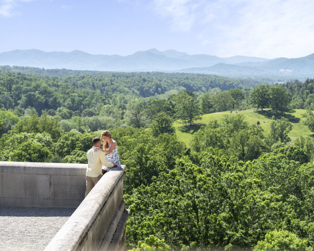 landscape image with newly engaged couple looking at each other and smiling while she sits on the ledge of the south terrace at biltmore estate with moutains in background