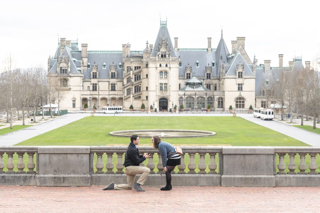couple standing on esplanade, man on one knee proposing, woman bent over happily surprised with Biltmore Estate in the background