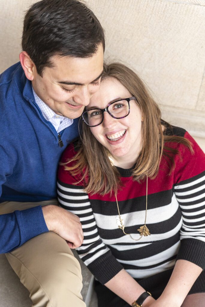 newly engaged couple sitting on staris. woman's back is leaned into fiance. she is smiling and looking at the camera, he is smiling and looking at her