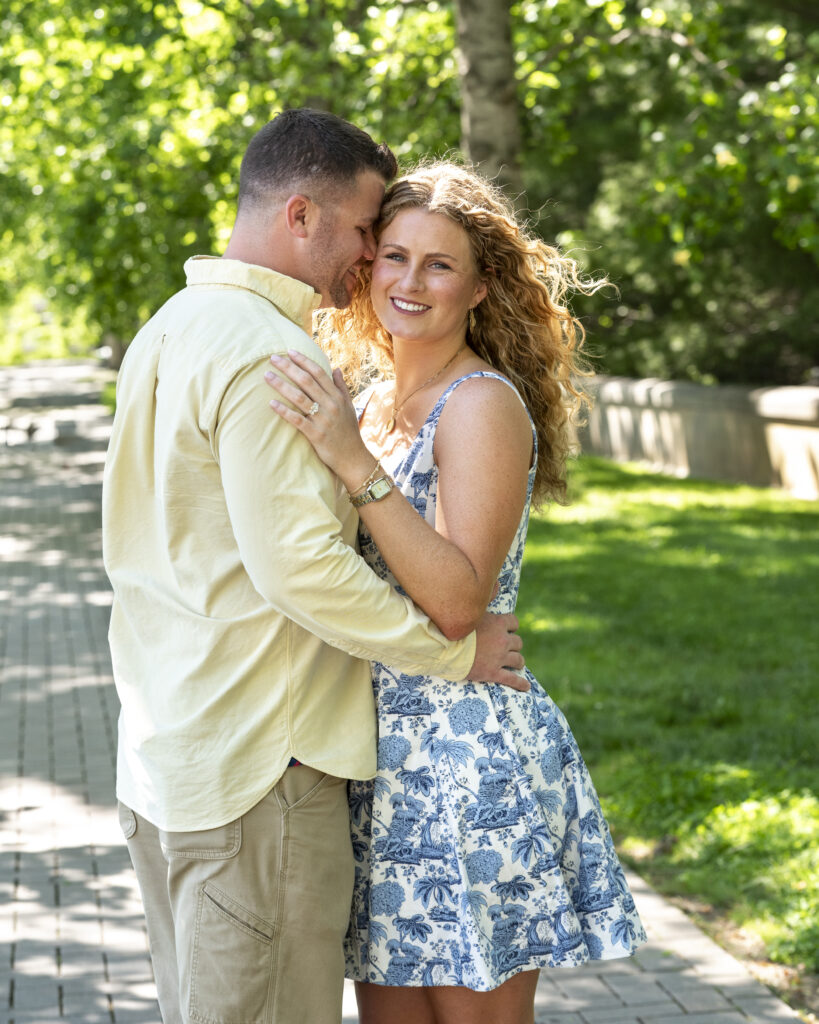 woman looking at camera smiling and fiance is kissing her temple on tree lined walking path in front of biltmore estate