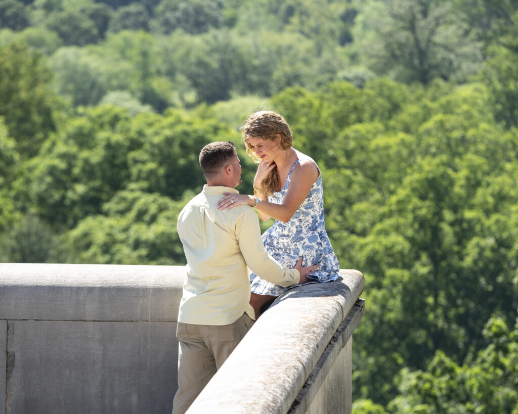 newly engaged couple looking at each other and smiling while she sits on the ledge of the south terrace at biltmore estate