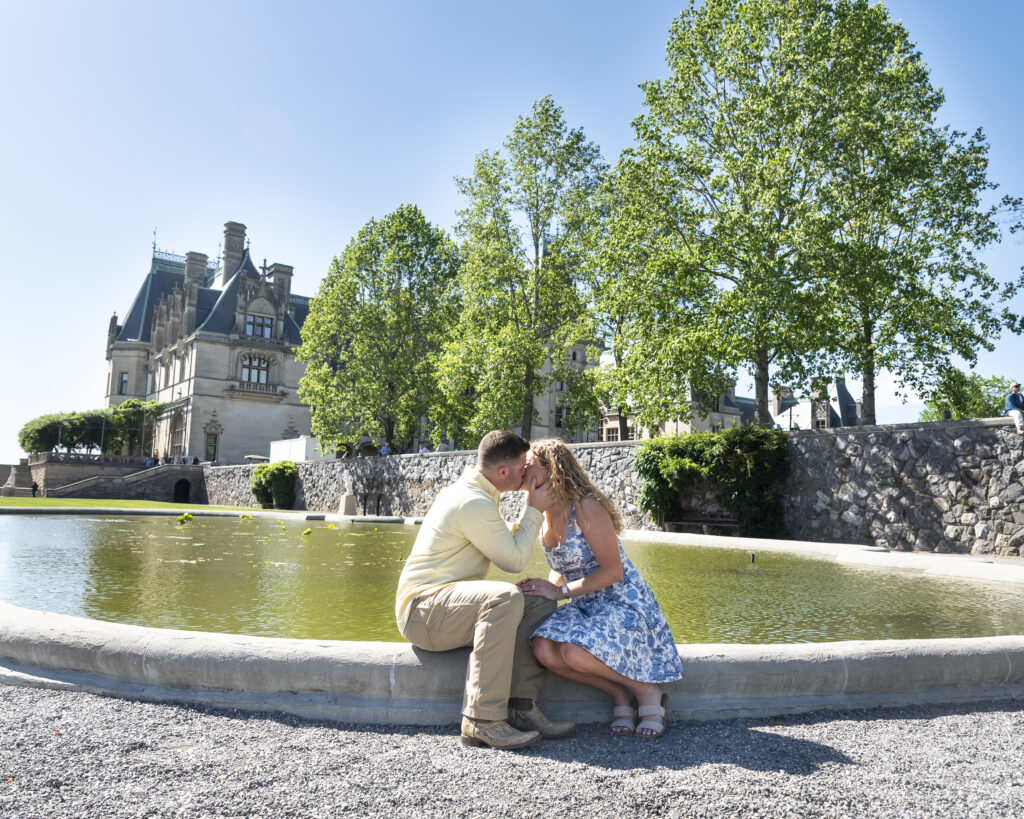 newly engaged couple sitting on the side of the coy pond kissing with Biltmore Estate in background on a sunny day