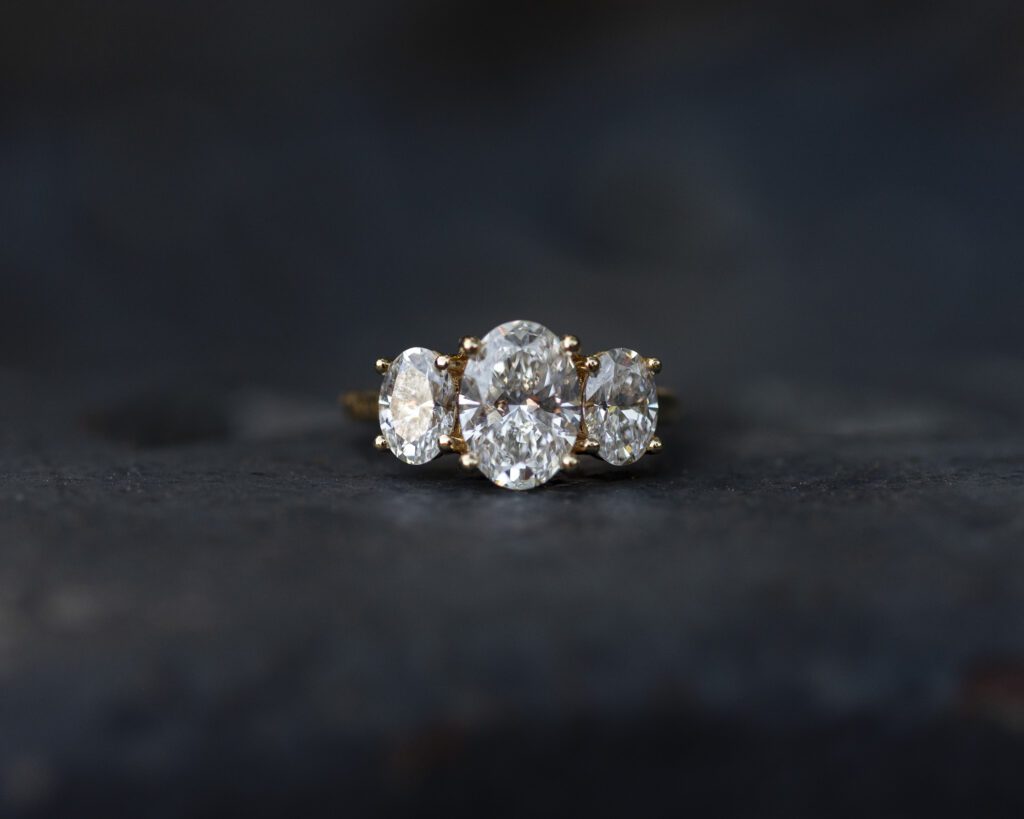 3 stone oval diamond and gold band engagement ring. blurry black background lets the ring stand out. used to proposal at Biltmore Estate