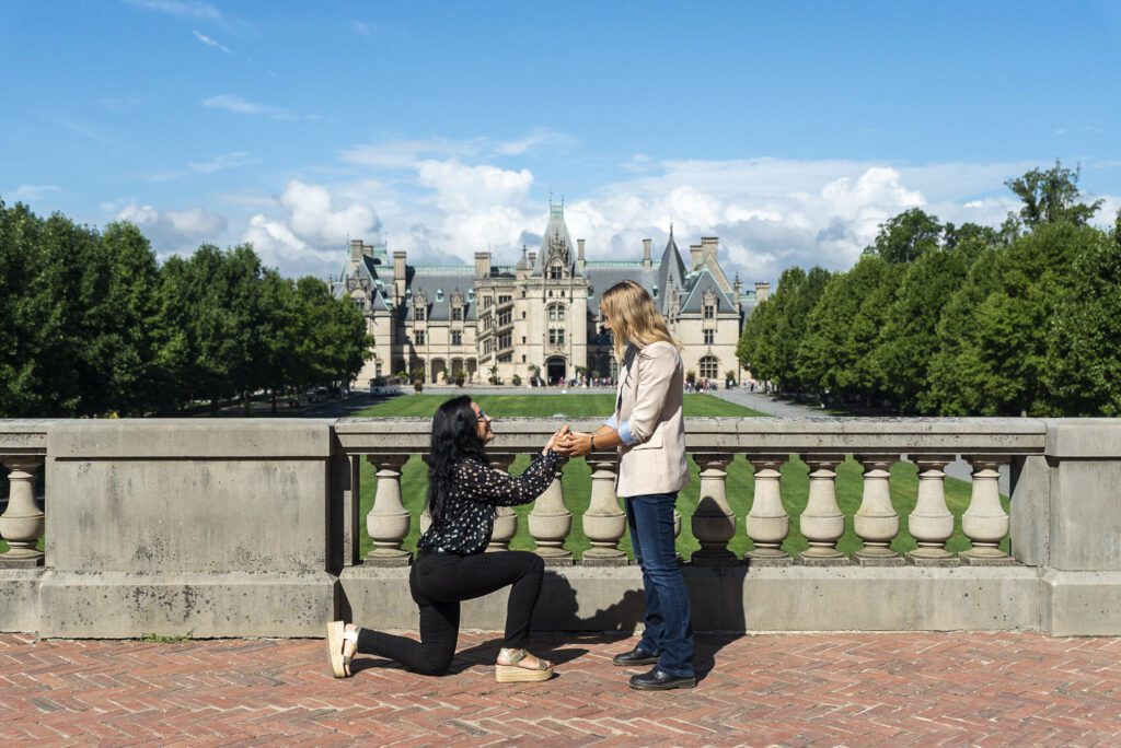 Biltmore estate proposal, lesbian couple, woman on one knee proposing to her girlfriend on the esplanade with the estate in the background on a sunny day