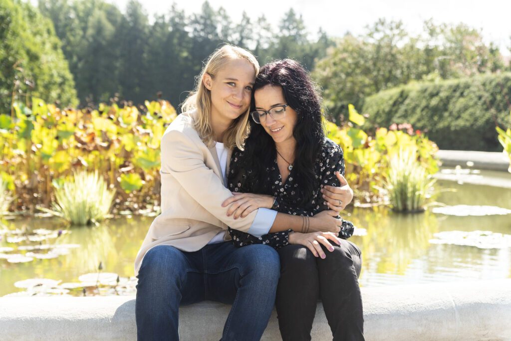 lesbian couple sitting on the side of the coy pond. One is looking at her ring, the other is looking at the camera on a sunny day