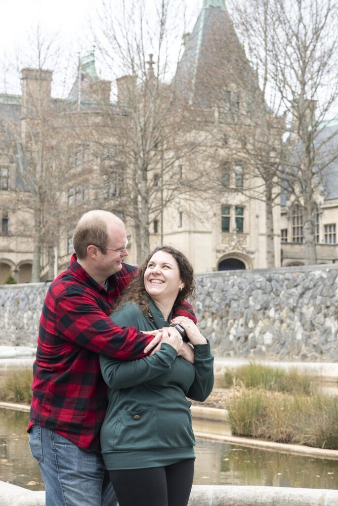 couple in sweaters embracing each other in Biltmore estate Italian gardens with the castle in the background after a proposal
