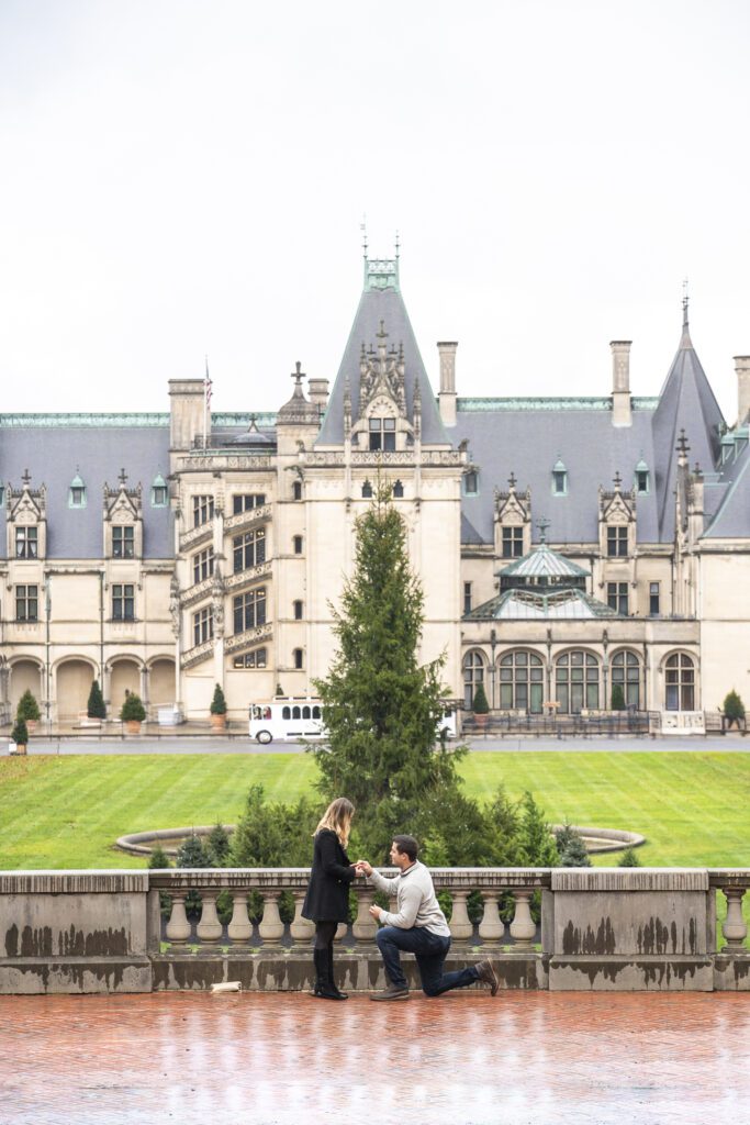 rainy day at Biltmore Estate. couple standing on esplanade, man on one knee proposing, woman standing happily surprised with Biltmore Estate and Christmas tree in the background