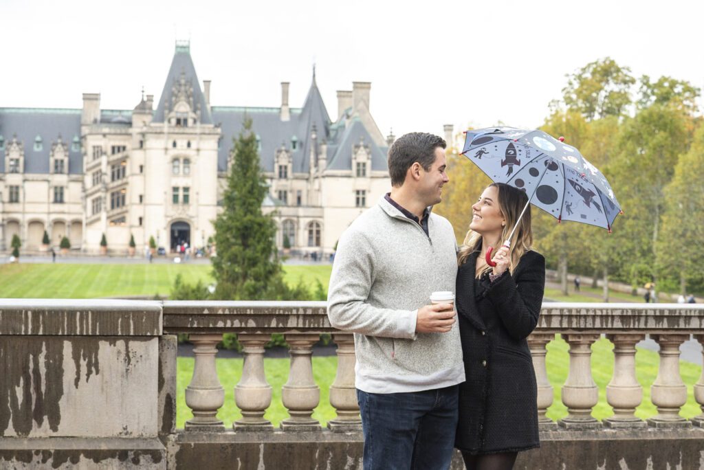 couple standing on esplanade after rainfall, couple looking at each other smiling while he holds a coffee and she holds an umbrella immediately after their proposal with Biltmore Estate in the background