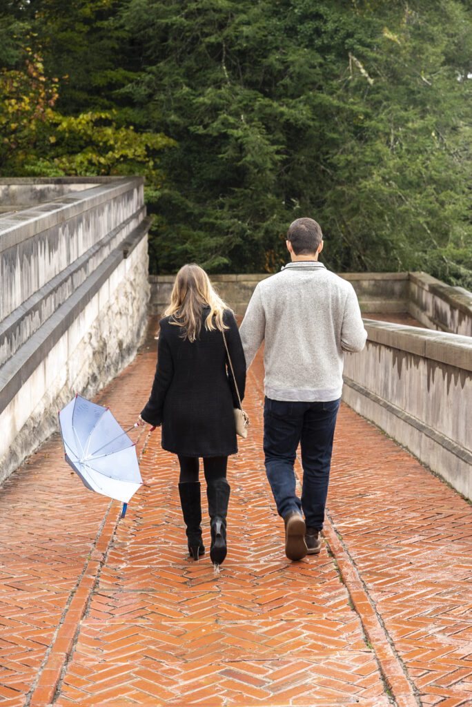 couple walking down path leaving the esplanade at Biltmore Estate after rainfall. couple looking forward while he holds a coffee and she holds a blue kids space umbrella immediately after their proposal