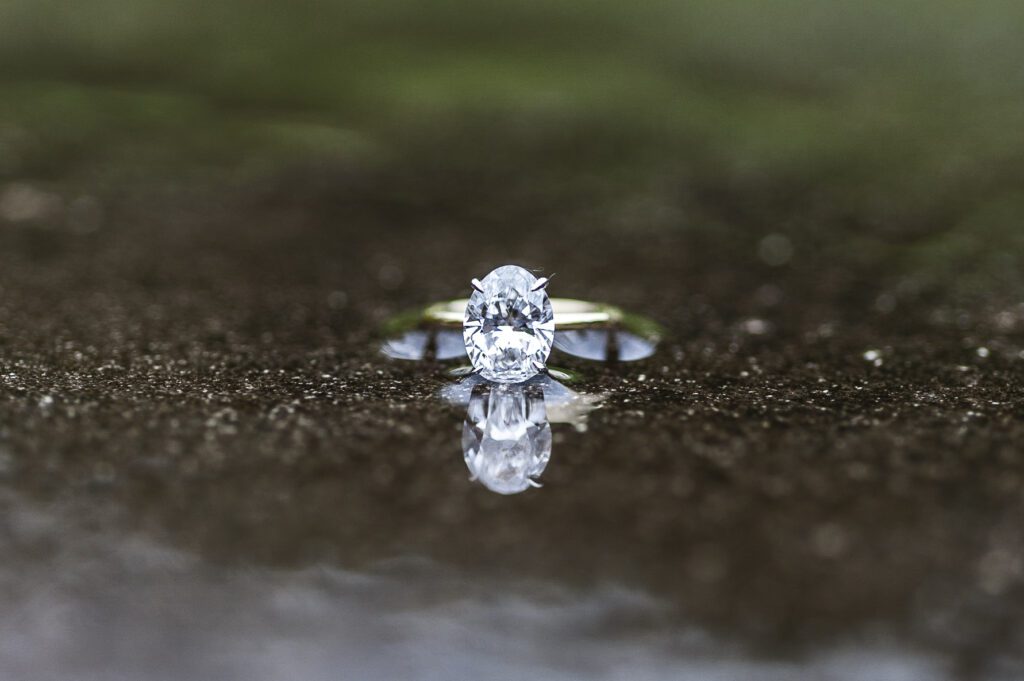 oval solitaire diamond ring in gold band sitting on wet concrete with a reflection from a puddle