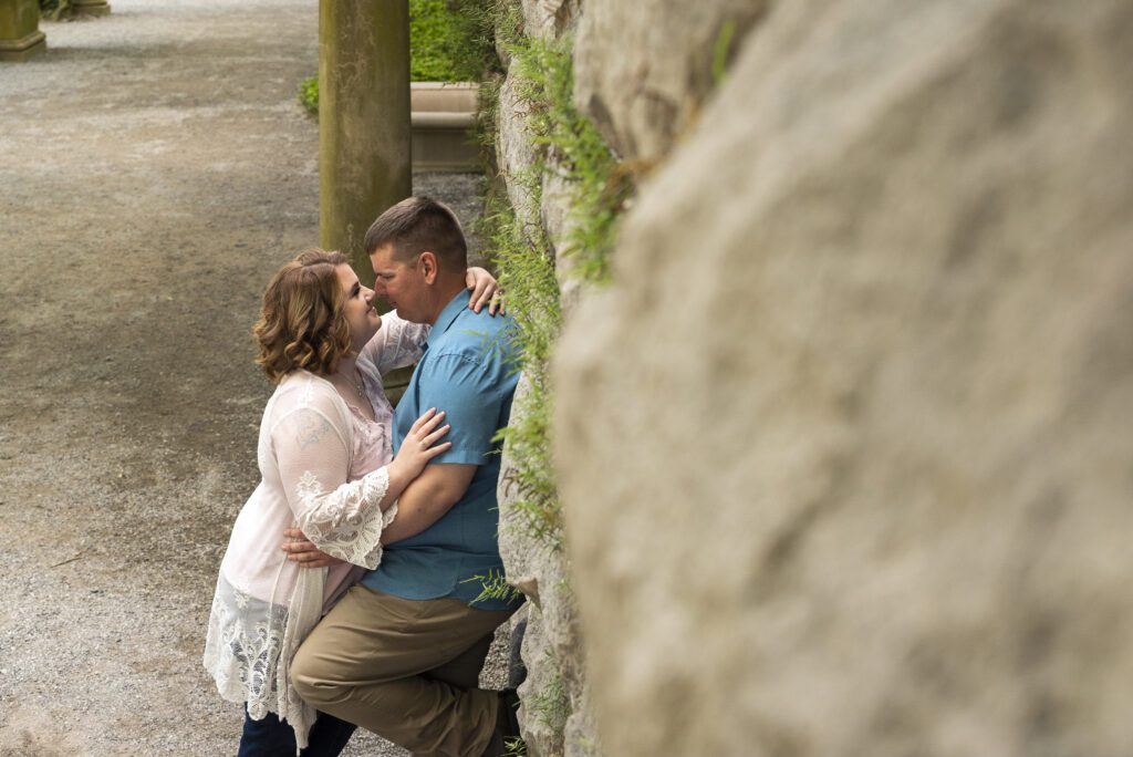man leaning on rock wall of whisteria garden at Biltmore estate, his fiance is leaning into him. embracing in an almost kiss moment. 