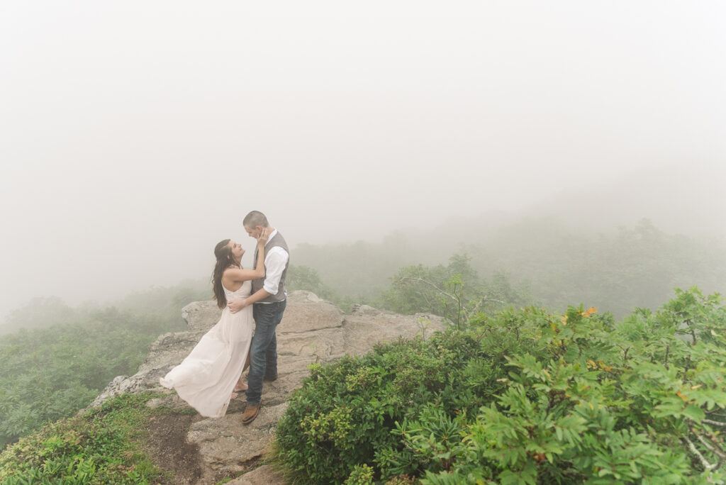 wide image of a couple standing on a rock at the top of Craggy gardens surrounded by fog while looking at each other and her dress flowing in the wind