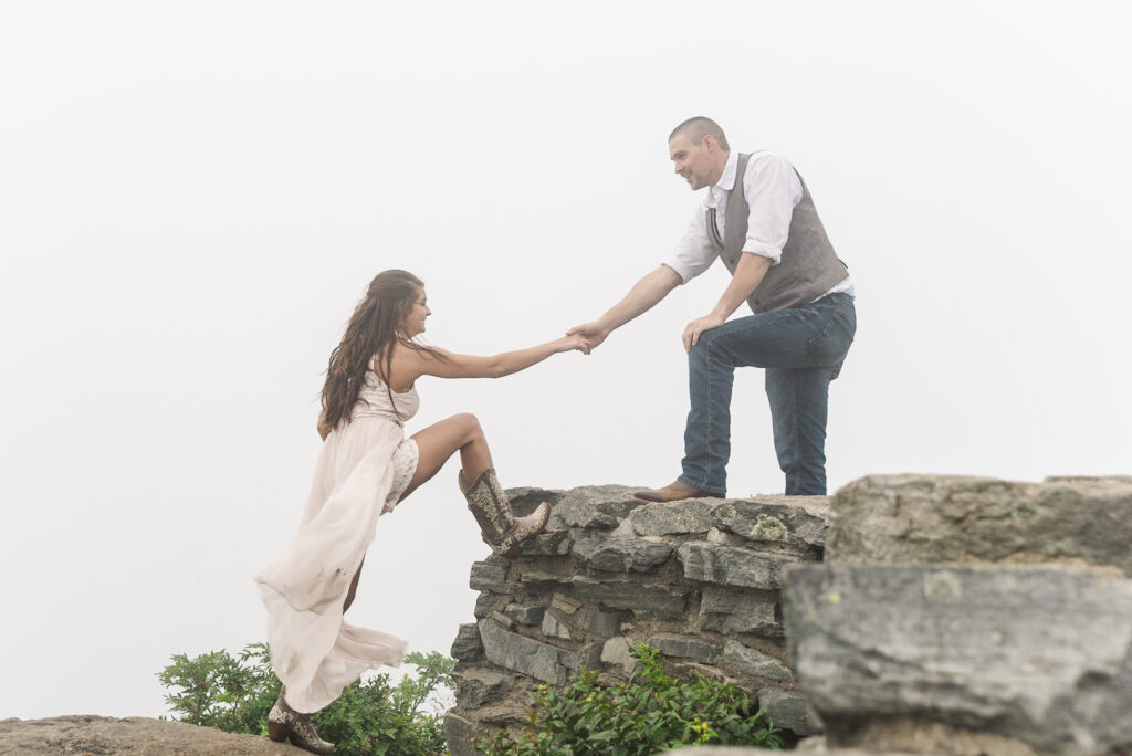 wide of a man reaching out to help his fiance step up on a rock as she reaches back for him