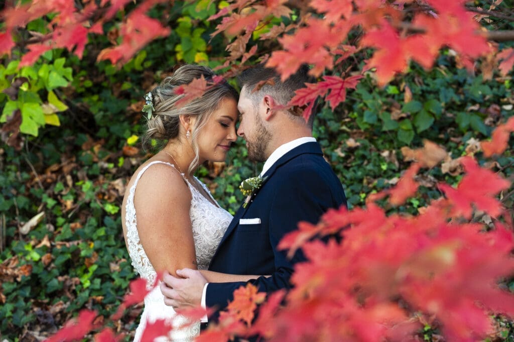 bride and groom embracing forehead to forehead during first look with red fall leaves framing them in foreground in asheville for crest center wedding