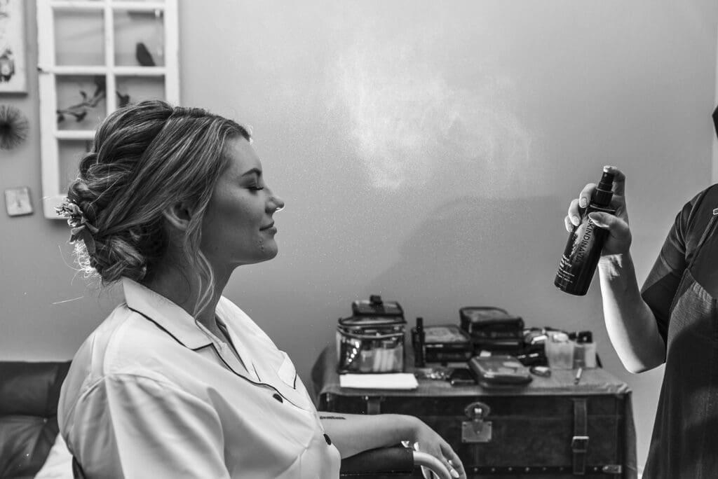 bride sitting in chair during hair and makeup as MUA applies setting spray