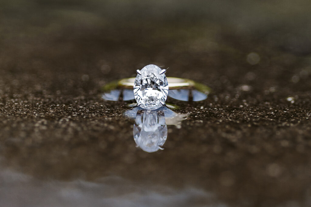 oval solitaire diamond ring in gold band sitting on wet concrete with a reflection from a puddle