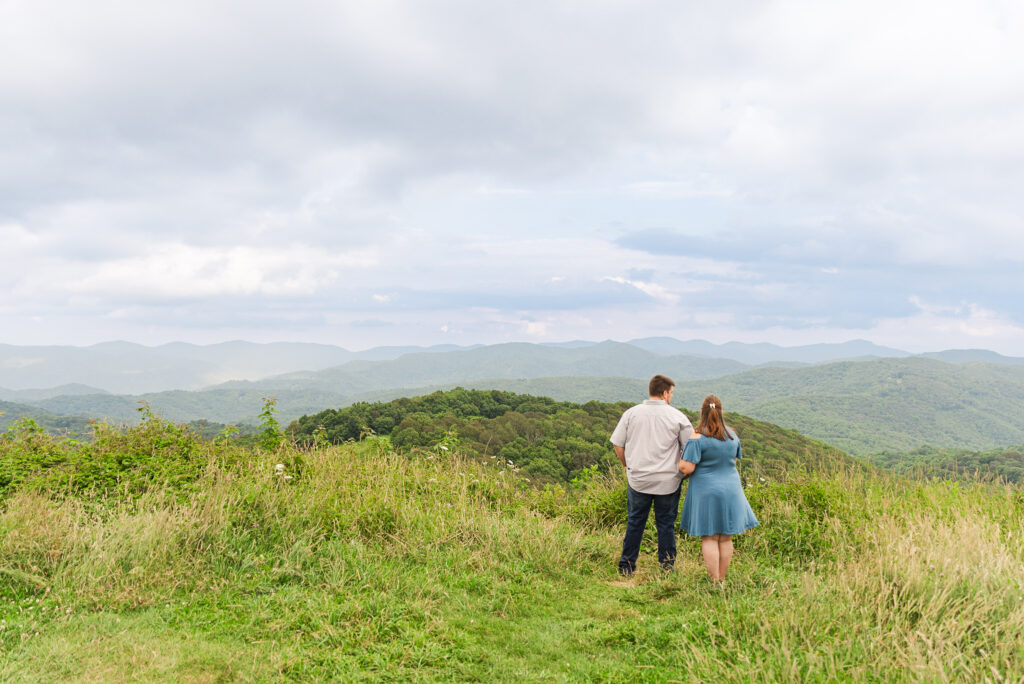 wide landscape image of max patch with engaged couple standing next to each other holding hands looking at the moubtains