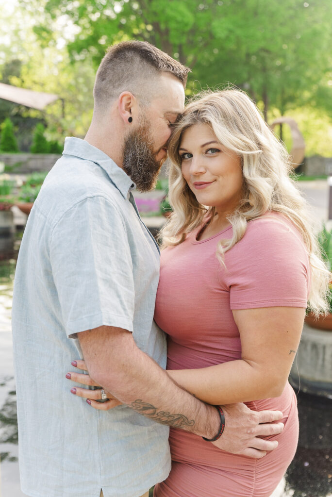 close up of couple standing chest to chest, she is looking at camera, he is looking at her as they stand in the NC arboretum 