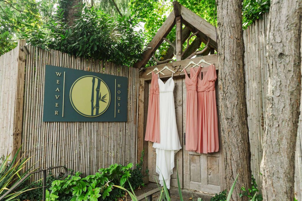 wide image of Weaver House wedding venue sign on the left with bride's and bridesmaid's dresses hanging on venue entrance gate on the right